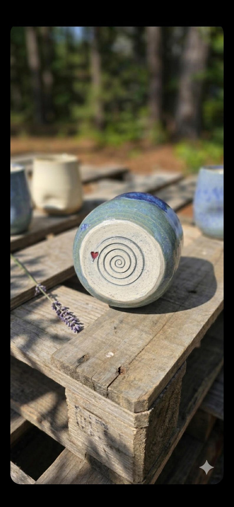 Puede incluir: Un jarr&oacute;n de cer&aacute;mica hecho a mano con un esmalte azul y verde, con un dise&ntilde;o en espiral y un peque&ntilde;o coraz&oacute;n rojo en la base. El jarr&oacute;n se exhibe sobre una superficie de madera r&uacute;stica, con otras piezas de cer&aacute;mica al fondo.