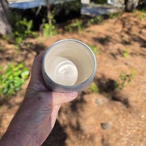 Puede incluir: Una taza de cer&aacute;mica hecha a mano con un exterior azul claro y un interior blanco. La taza se sostiene en una mano contra un fondo borroso al aire libre. El borde de la taza tiene un sutil esmalte azul. La taza parece estar vac&iacute;a.