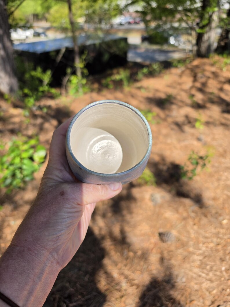 Puede incluir: Una taza de cer&aacute;mica hecha a mano con un exterior azul claro y un interior blanco. La taza se sostiene en una mano contra un fondo borroso al aire libre. El borde de la taza tiene un sutil esmalte azul. La taza parece estar vac&iacute;a.