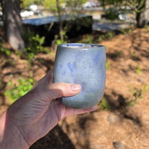Puede incluir: Un vaso de cer&aacute;mica hecho a mano con un esmalte azul moteado. La taza tiene una forma redondeada y se sostiene en una mano, con un fondo borroso al aire libre de &aacute;rboles y luz solar.