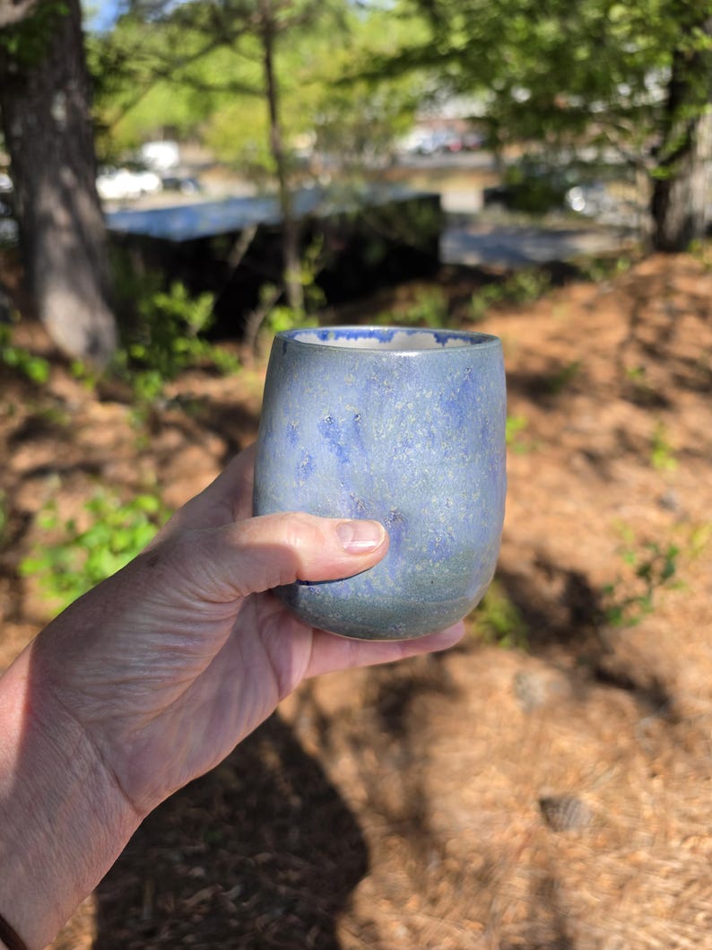 Puede incluir: Un vaso de cer&aacute;mica hecho a mano con un esmalte azul moteado. La taza tiene una forma redondeada y se sostiene en una mano, con un fondo borroso al aire libre de &aacute;rboles y luz solar.