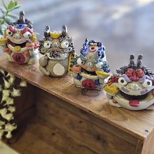 May include: Four colorful ceramic figurines of mythical creatures with expressive faces and intricate details. Each figure has unique features, including horns, and holds objects. They are arranged on a wooden surface.