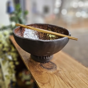 Op de afbeelding: Een donkerbruine keramische kom met een textuur aan de binnenkant en een decoratieve voet. Een paar bamboe eetstokjes rust over de rand van de kom. De kom staat op een houten oppervlak.