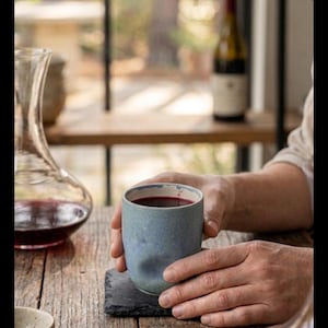 Puede incluir: Una persona sosteniendo una taza de cer&aacute;mica azul llena de l&iacute;quido rojo, probablemente vino, sobre una mesa de madera. Una jarra de vino y una botella de vino est&aacute;n en el fondo. Un corcho y un cuenco peque&ntilde;o tambi&eacute;n est&aacute;n sobre la mesa.