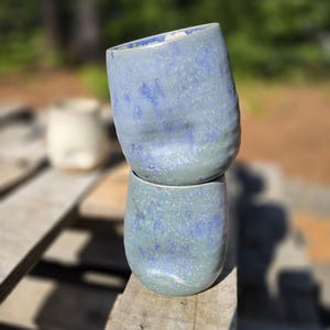Puede incluir: Dos tazas de cer&aacute;mica azul hechas a mano apiladas sobre una superficie de madera. Las tazas tienen un aspecto texturizado y moteado con tonos de azul y verde. Las tazas est&aacute;n ligeramente inclinadas, mostrando su forma org&aacute;nica &uacute;nica. El fondo est&aacute; borroso, con toques de follaje verde.