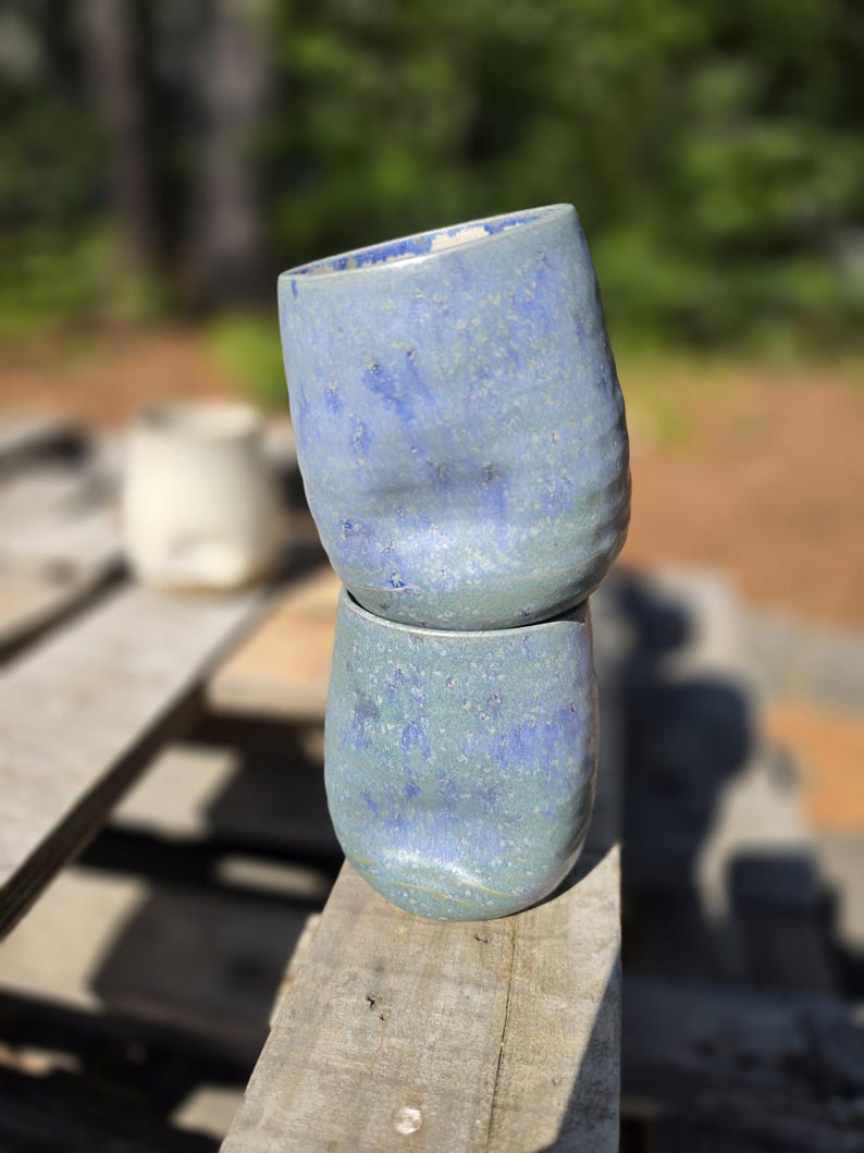 Puede incluir: Dos tazas de cer&aacute;mica azul hechas a mano apiladas sobre una superficie de madera. Las tazas tienen un aspecto texturizado y moteado con tonos de azul y verde. Las tazas est&aacute;n ligeramente inclinadas, mostrando su forma org&aacute;nica &uacute;nica. El fondo est&aacute; borroso, con toques de follaje verde.