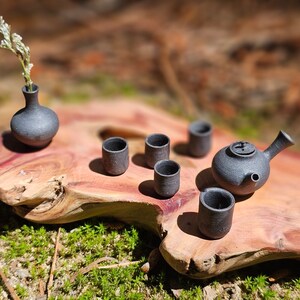 Może przedstawiać: Miniaturowy czarny ceramiczny zestaw do herbaty, w tym czajniczek, filiżanki i mały wazon z gałązką kwiatów. Zestaw jest prezentowany na rustykalnej drewnianej płycie z rozmytym naturalnym tłem.