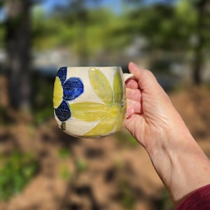 May include: A ceramic mug with a cream base, decorated with large, stylised floral designs in yellow and blue. The mug has a rounded shape with a handle. It is held in a person's hand.