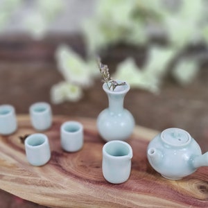 Könnte beinhalten: Ein hellblaues Keramik-Teeservice, bestehend aus einer Teekanne, sechs kleinen Tassen und einer kleinen Vase mit getrockneten Blumen. Das Set wird auf einer Holzoberfläche präsentiert und erzeugt eine ruhige und minimalistische Ästhetik.