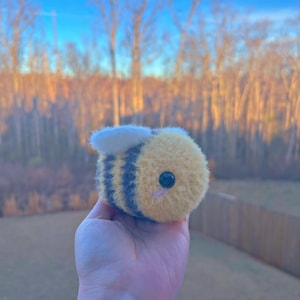 May include: A plush bee toy with a yellow body, black stripes, and a white wing. The bee has a small pink mouth and a black eye. The toy is held in a hand against a blurred outdoor background.
