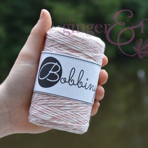 May include: A spool of pink and white twine with a label that reads "Bobbin". The twine is wrapped around a cardboard core. The image is taken in natural light, with a blurred background.