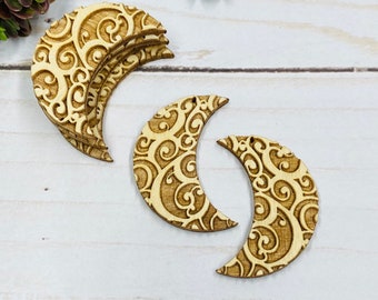 May include: Light brown wooden crescent moon shapes with an engraved swirling pattern. The moons have a small hole for hanging. Several are stacked, with others lying separately on a white wooden surface. Ideal for crafts.