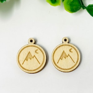 May include: Two wooden circle pendants featuring a mountain and crescent moon design. The pendants are light brown and have a hole at the top for hanging.