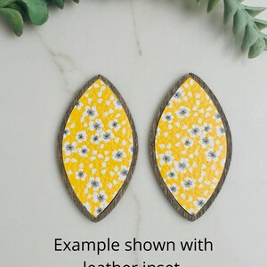 May include: Two teardrop earrings with a yellow floral pattern and a dark brown wooden frame. The earrings are shown on a white surface with a green sprig. The text "Example shown with leather inset" is visible.