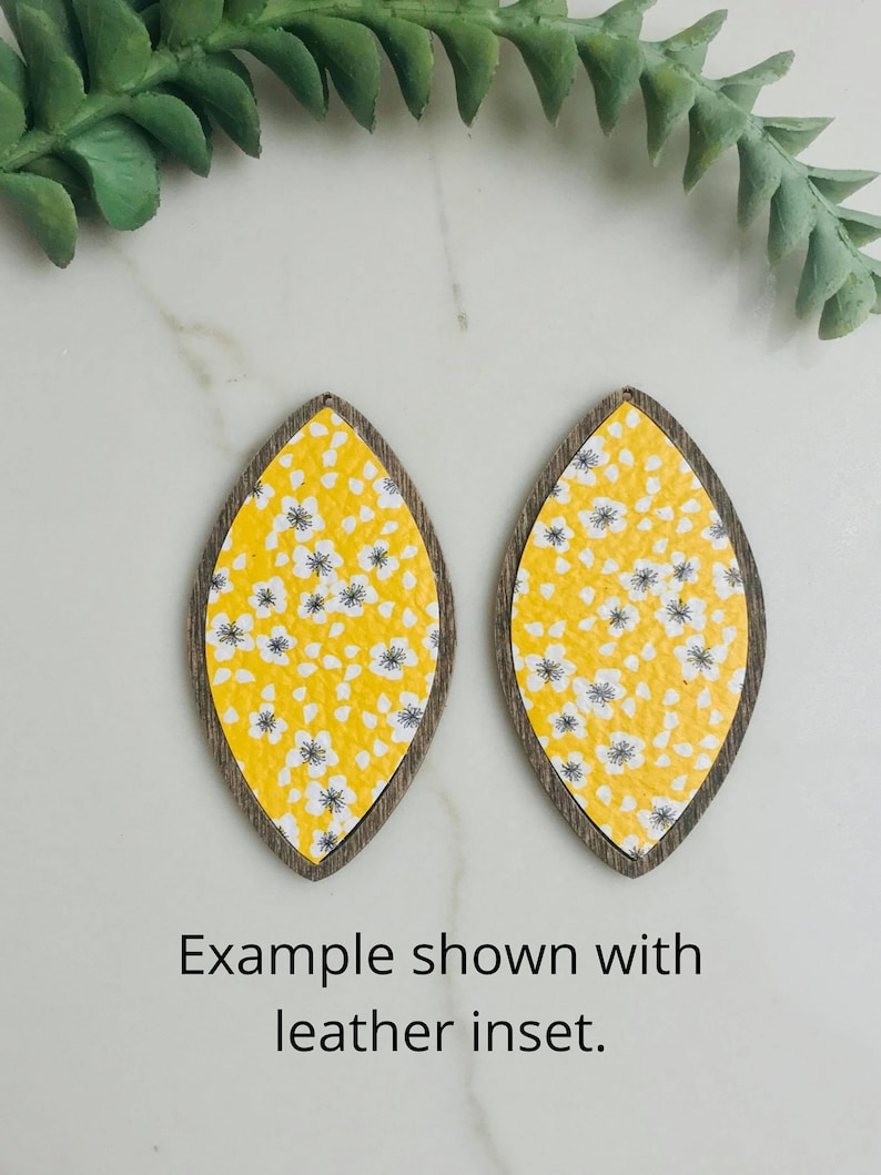 May include: Two teardrop earrings with a yellow floral pattern and a dark brown wooden frame. The earrings are shown on a white surface with a green sprig. The text "Example shown with leather inset" is visible.