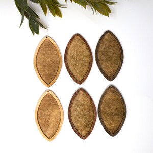 May include: Six wooden teardrop-shaped earrings in varying shades of brown. The earrings have a light-coloured centre and a darker, contrasting border. Each earring has a small hole at the top for hanging. Green leaves are in the upper left corner.