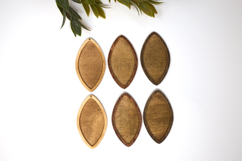 May include: Six wooden teardrop-shaped earrings in varying shades of brown. The earrings have a light-coloured centre and a darker, contrasting border. Each earring has a small hole at the top for hanging. Green leaves are in the upper left corner.