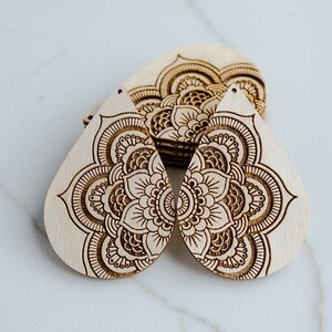May include: Two wooden teardrop-shaped earrings with a laser-etched mandala design. The earrings are light brown and have a natural wood finish.
