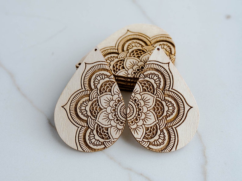 May include: Two wooden teardrop-shaped earrings with a laser-etched mandala design. The earrings are light brown and have a natural wood finish.