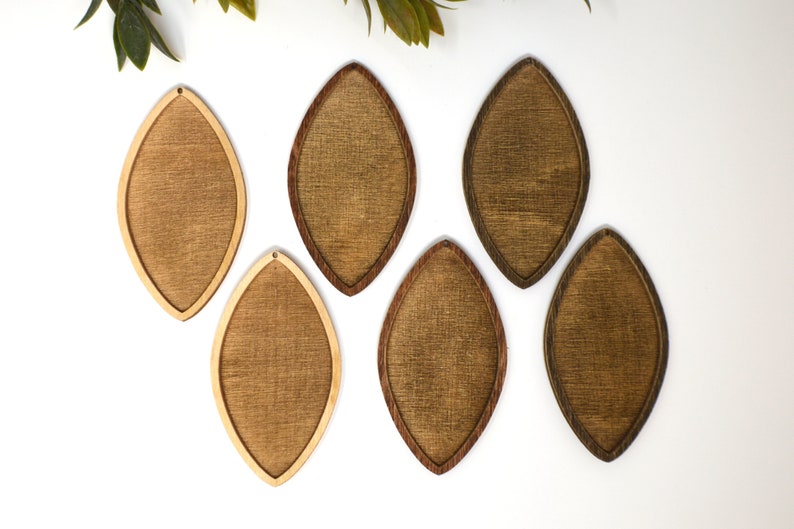 May include: Six wooden teardrop-shaped earrings in varying shades of brown. The earrings have a light-coloured wood grain centre with a darker wood border. Each earring has a small hole at the top for hanging. The earrings are displayed on a white surface.