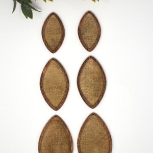 May include: Six wooden teardrop-shaped earrings. Each earring has a textured, light brown centre and a dark brown border. They are arranged on a white surface, with green leaves in the upper left corner.