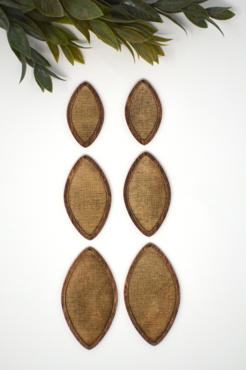 May include: Six wooden teardrop-shaped earrings. Each earring has a textured, light brown centre and a dark brown border. They are arranged on a white surface, with green leaves in the upper left corner.