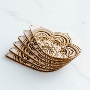 May include: A stack of six unfinished wooden cutouts with intricate mandala designs. The cutouts are in the shape of a teardrop and have a natural wood color.