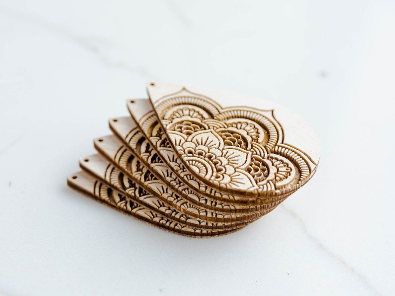 May include: A stack of six unfinished wooden cutouts with intricate mandala designs. The cutouts are in the shape of a teardrop and have a natural wood color.