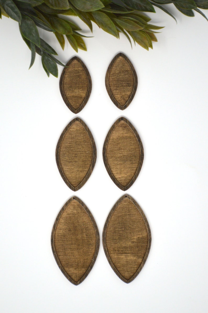 May include: Six wooden teardrop-shaped earring blanks with a dark brown stain and a lighter brown centre. The earrings are arranged on a white surface, with green leaves in the upper left corner.