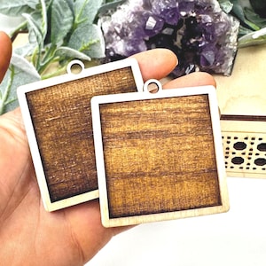 May include: Two square wooden pendants with a light wood frame and a dark wood grain center. Each pendant has a small hole at the top for hanging. The pendants are held in a hand, with a wooden box visible in the background.