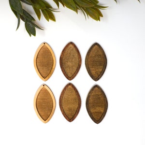 May include: Six wooden teardrop-shaped earrings in varying shades of brown. The earrings have a lighter wood centre and a darker wood border. The earrings are arranged on a white surface, with green leaves in the upper left corner.