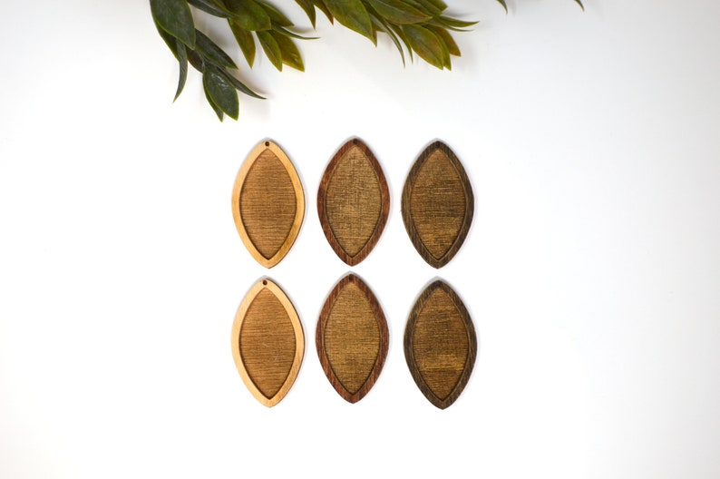 May include: Six wooden teardrop-shaped earrings in varying shades of brown. The earrings have a lighter wood centre and a darker wood border. The earrings are arranged on a white surface, with green leaves in the upper left corner.