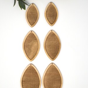 May include: Six unfinished wooden teardrop-shaped earring blanks with a textured centre and a smooth, light-coloured border. Each blank has a small hole at the top for attaching earring hooks. Green leaves are in the upper left corner.