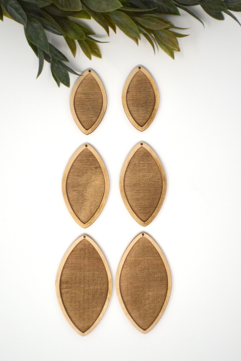 May include: Six unfinished wooden teardrop-shaped earring blanks with a textured centre and a smooth, light-coloured border. Each blank has a small hole at the top for attaching earring hooks. Green leaves are in the upper left corner.