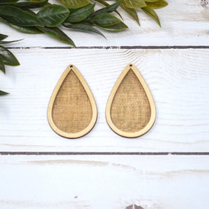 May include: Two unfinished wooden teardrop-shaped earring blanks. The blanks have a light brown wood grain texture within a lighter wood border. The earrings are shown on a white wooden surface with green leaves in the background.