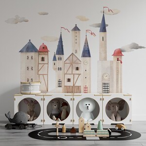 Może przedstawiać: Biały drewniany domek do zabawy z trzema okrągłymi otworami, w których znajdują się pluszowe zabawki. Domek do zabawy jest ozdobiony fantazyjną akwarelą przedstawiającą zamek z czerwonymi i niebieskimi akcentami. Czarno-biały tor wyścigowy znajduje się na podłodze przed domkiem do zabawy.