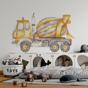 Puede incluir: Una ilustración de acuarela de un camión mezclador de cemento amarillo y gris en una pared blanca. El camión está sobre un estante blanco con una cesta de almacenamiento de juguetes etiquetada como "TOYS" y bloques de madera con letras.