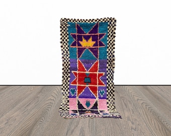 May include: A colorful Moroccan rug with a geometric pattern featuring stars and diamonds in shades of red, blue, purple, pink, and yellow. The rug has a black and white checkered border.