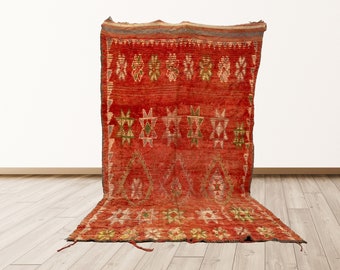Puede incluir: Una alfombra roja marroquí con un patrón geométrico en tonos de verde, amarillo y blanco. La alfombra tiene un borde con flecos y está tejida con un diseño tradicional.