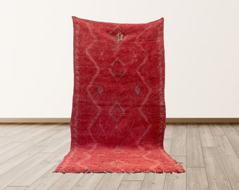 May include: A red Moroccan rug with a diamond pattern. The rug has a fringed edge and is made of wool.