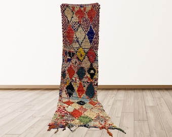 May include: A long, colorful Moroccan rug with a diamond pattern in shades of red, blue, green, yellow, and white. The rug has a fringe on the bottom and is made of wool.