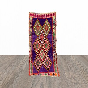 May include: A long, narrow rug with a purple background and a colourful diamond pattern. The rug has a fringe border and is made of wool.