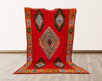 May include: A red Moroccan rug with a geometric pattern in shades of orange, green, blue, and yellow. The rug has a diamond-shaped center with smaller diamond shapes within it. The rug is surrounded by a border of stars and other geometric shapes.