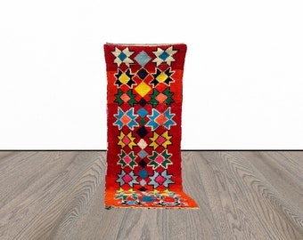 May include: A colorful, geometric patterned rug with a red background. The rug has a long, narrow shape and features a variety of colors, including blue, green, yellow, and white.
