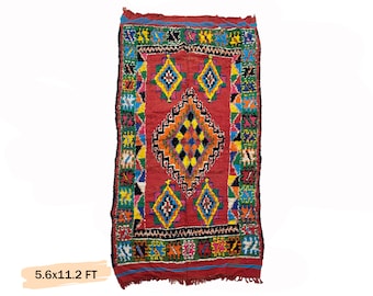 May include: A colorful Moroccan rug with a red background and intricate geometric patterns in shades of blue, green, yellow, and black. The rug measures 5.6 x 11.2 feet.