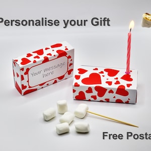 May include: Two white boxes with red hearts printed on them. One box has a blank label that says "Your message here". The other box has a pink and white striped candle in it. There are white marshmallows and wooden skewers on a white surface. The text "Free Postage" is printed on the surface.