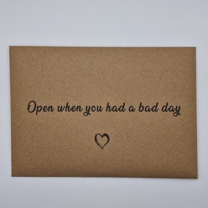 K&ouml;nnte beinhalten: Ein brauner Papierumschlag mit dem Text "Open when you had a bad day" und einem kleinen gezeichneten Herzen.