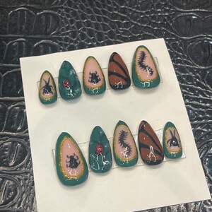 Könnte beinhalten: Ein Set aus zehn künstlichen Nägeln mit verschiedenen Designs. Jeder Nagel hat eine dunkelgrüne Basis mit goldfarbenem Glitzerrand. Designs umfassen Insekten, florale Elemente und abstrakte Muster in Braun, Rosa und Weiß.