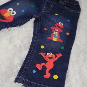 May include: Blue denim jeans with colorful polka dots and Elmo appliques. Elmo is holding blocks that spell "ELMO".