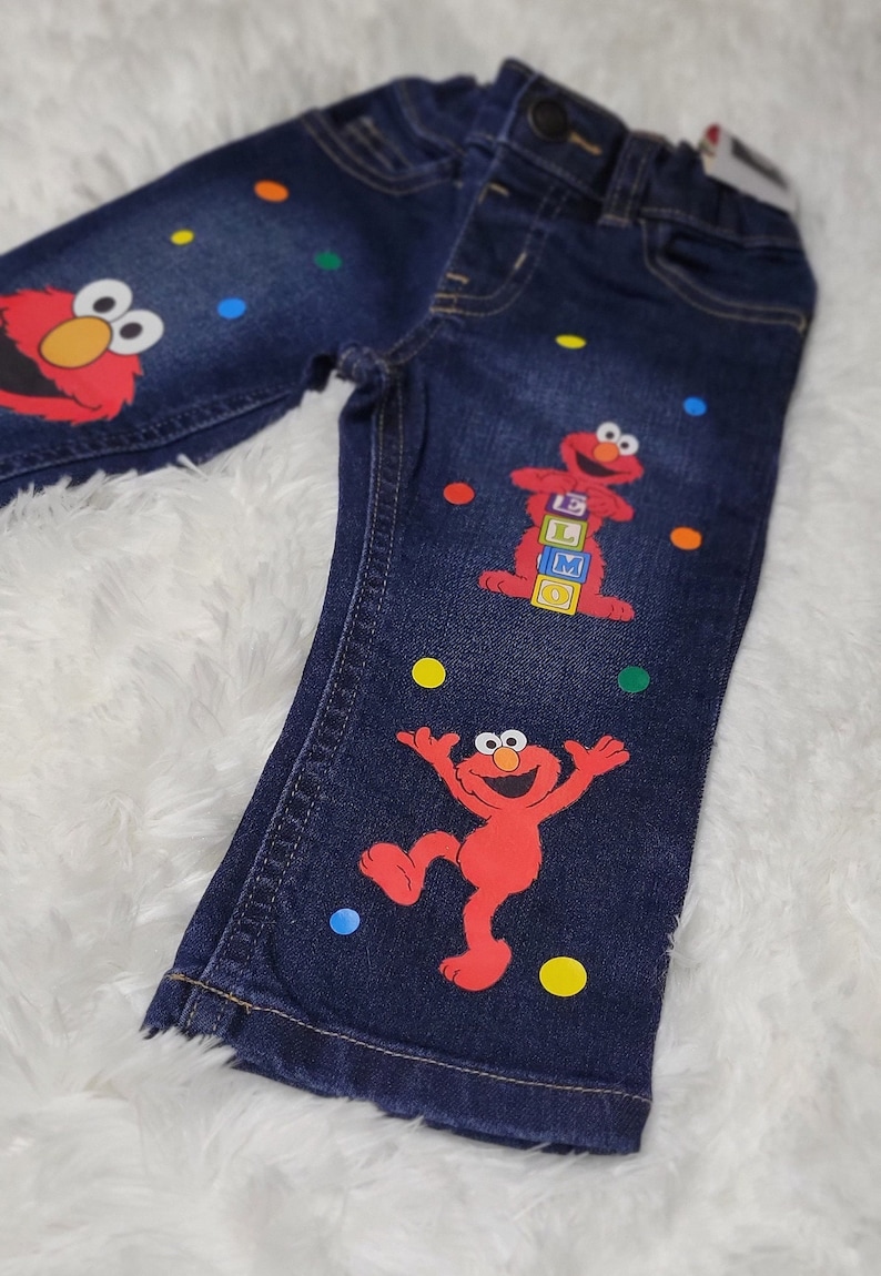 May include: Blue denim jeans with colorful polka dots and Elmo appliques. Elmo is holding blocks that spell "ELMO".